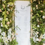 A Custom Printed Wedding Table Plan for Elizabeth and Larry features lush green foliage, white and peach flowers, and candlelit glass holders, making an elegant and romantic display.