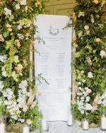 A Custom Printed Wedding Table Plan for Elizabeth and Larry features lush green foliage, white and peach flowers, and candlelit glass holders, making an elegant and romantic display.