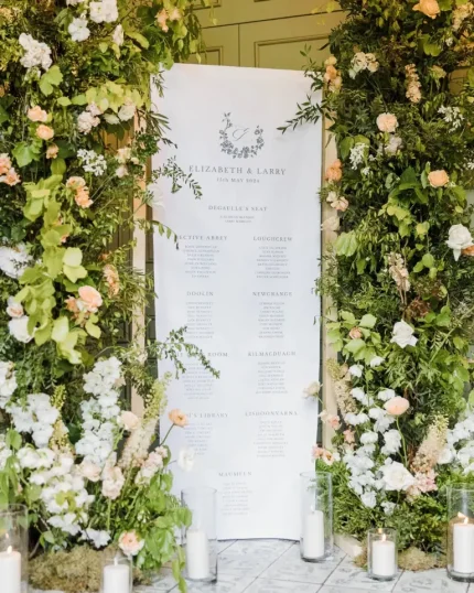 A Custom Printed Wedding Table Plan for Elizabeth and Larry features lush green foliage, white and peach flowers, and candlelit glass holders, making an elegant and romantic display.