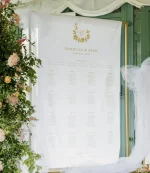 A Custom Printed Wedding Table Plan for Danielle & Seán (13th July 2024) is displayed near green doors with floral decor, tulle draping, and a personalised backdrop. Guest names appear under numbered table headings.