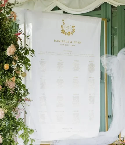 A Custom Printed Wedding Table Plan for Danielle & Seán (13th July 2024) is displayed near green doors with floral decor, tulle draping, and a personalised backdrop. Guest names appear under numbered table headings.