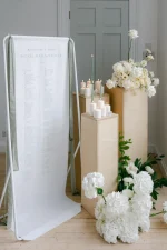 A Custom Printed Wedding Table Plan on white fabric stands by wooden pedestals with candles and white floral arrangements in a bright, elegant room with light wood floors.