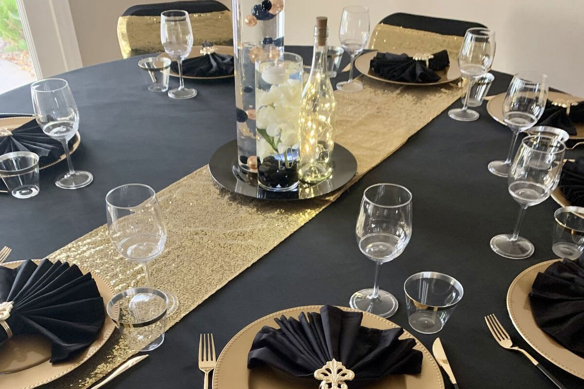 A round table set for a formal event, featuring personalised tablecloths or trade show table covers, gold sequin table runner, gold plates with black serviettes, wine glasses, and a decorative centrepiece with flowers and a bottle with fairy lights.