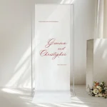 The Custom Printed Wedding Welcome Sign Banner features elegant red script on white, reading “Welcome to our wedding, Gemma and Christopher, 17 March 2023.” Displayed on a clear frame with flowers, it’s a perfect personalised backdrop for your big day.