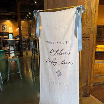 A Custom Printed Wedding Welcome Sign Banner with blue ribbons reads "Welcome to Chelsea’s baby shower, 10 August 2025," displayed in a rustic venue with wooden decor and metal bar stools.