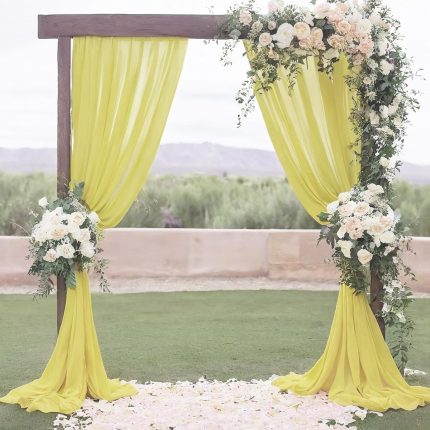 An ivory chiffon 4m x 4m stage backdrop drapes a wooden wedding arch adorned with white and peach flowers, set on grass with scattered petals and greenery in the background.