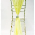 A rustic white wooden chair features the Chiffon Chiavari Vertical Drop Light Yellow—a sheer yellow sash gathered at the centre with a crystal brooch for elegant wedding chair décor, shown against a plain white background.